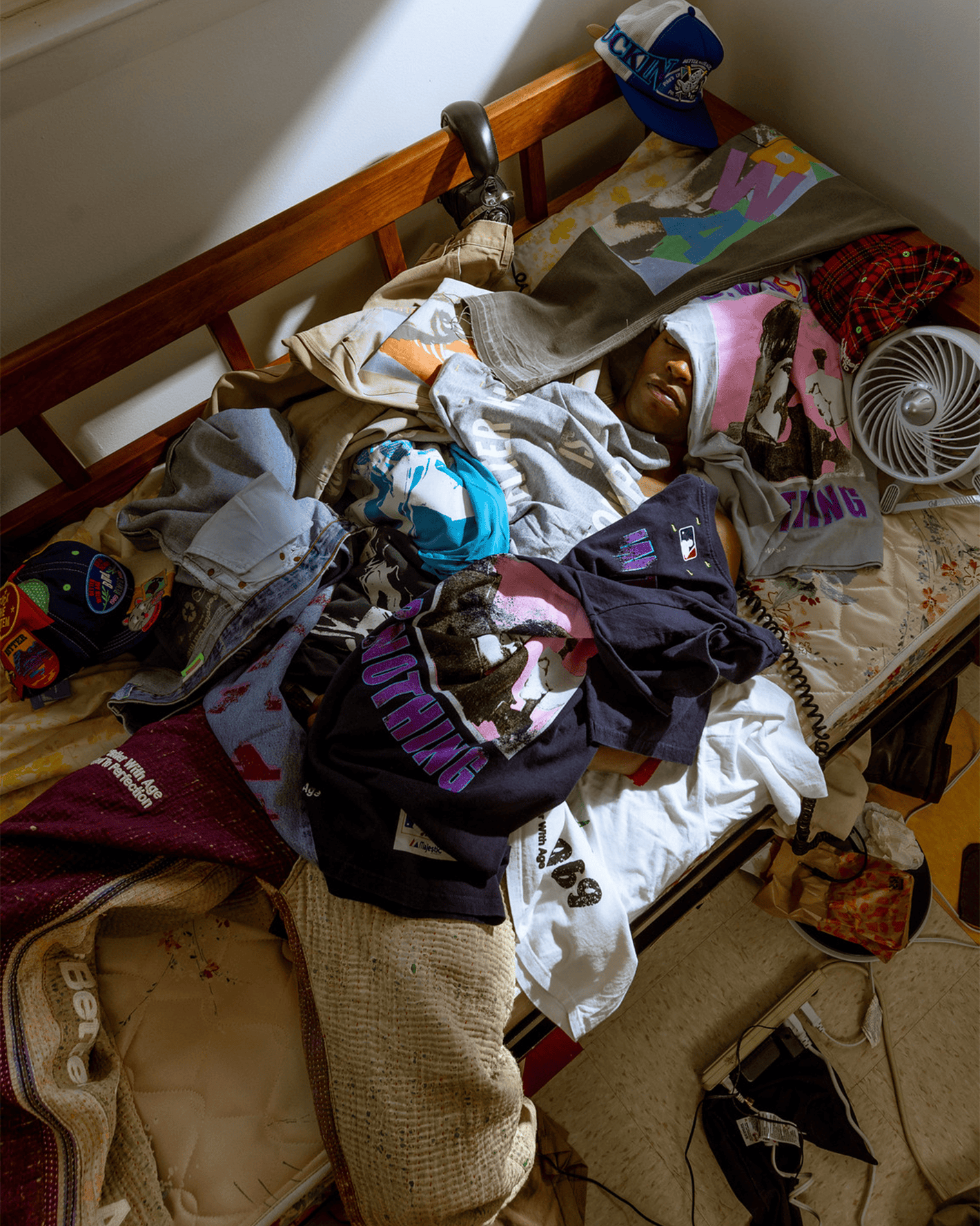 Person lying on a bed with clothes scattered around, in a room with a fan and various items.